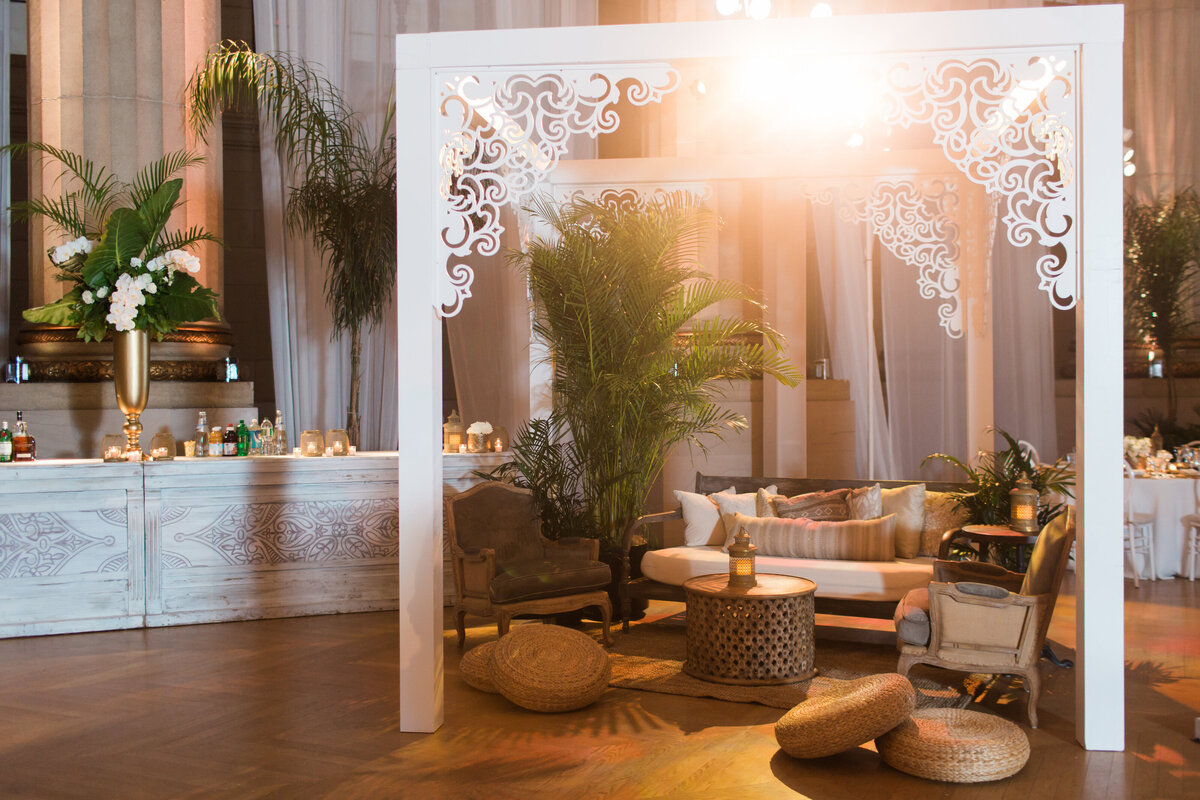 This cozy lounge area in an elegant event space features a white pergola with intricate designs, a plush sofa, chairs, and potted plants. The space is warmly lit, and nearby, a bar with floral arrangements sits against a backdrop of draped white curtains. This is where the photo was taken at Washington D.C., The Mellon Auditorium, one of the best luxury DC venues with European flair, captured beautifully by photographer Sarah Bradshaw.