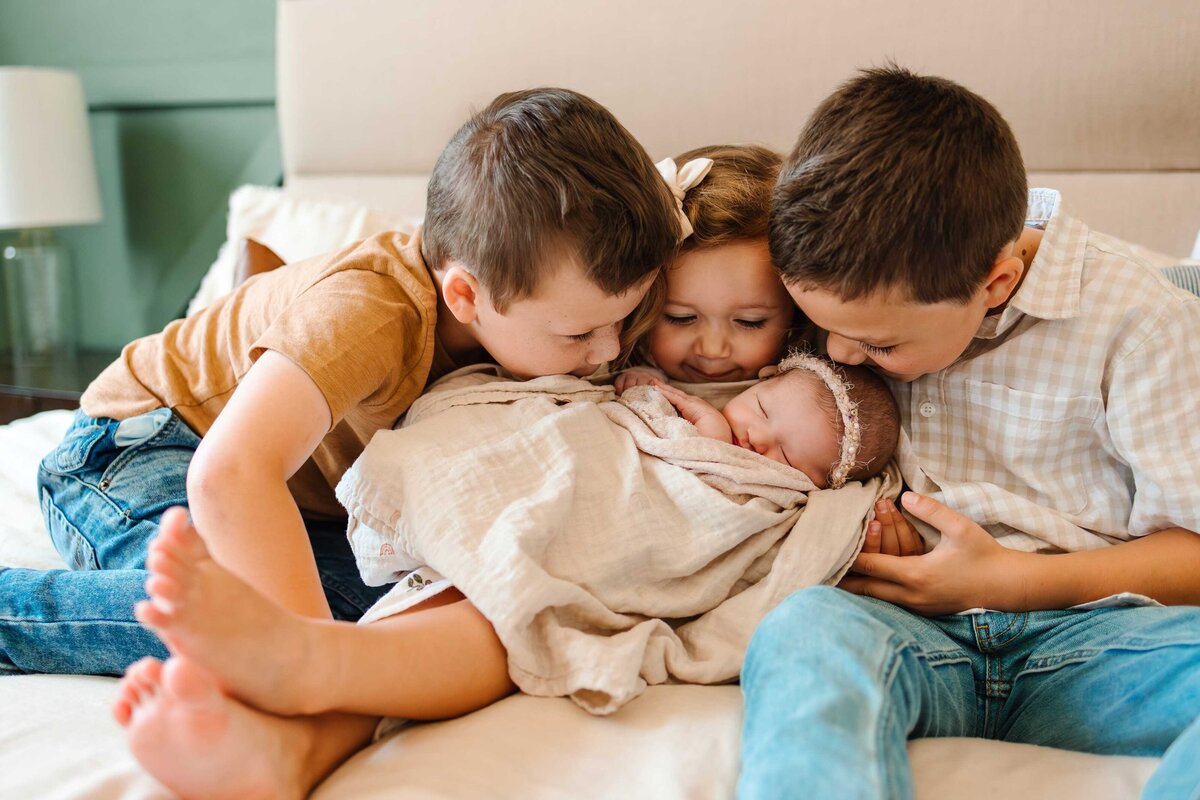 A heartwarming scene captured by a newborn photographer in Albuquerque. The newborn baby girl is peacefully sleeping, wrapped in a white blanket, while her three older siblings gaze at her with love and kindness, creating a tender family moment.