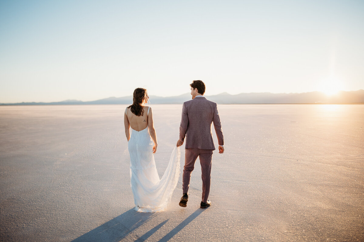 Bride and groom walking hand in hand across a vast salt flat, with the sunset casting a warm glow