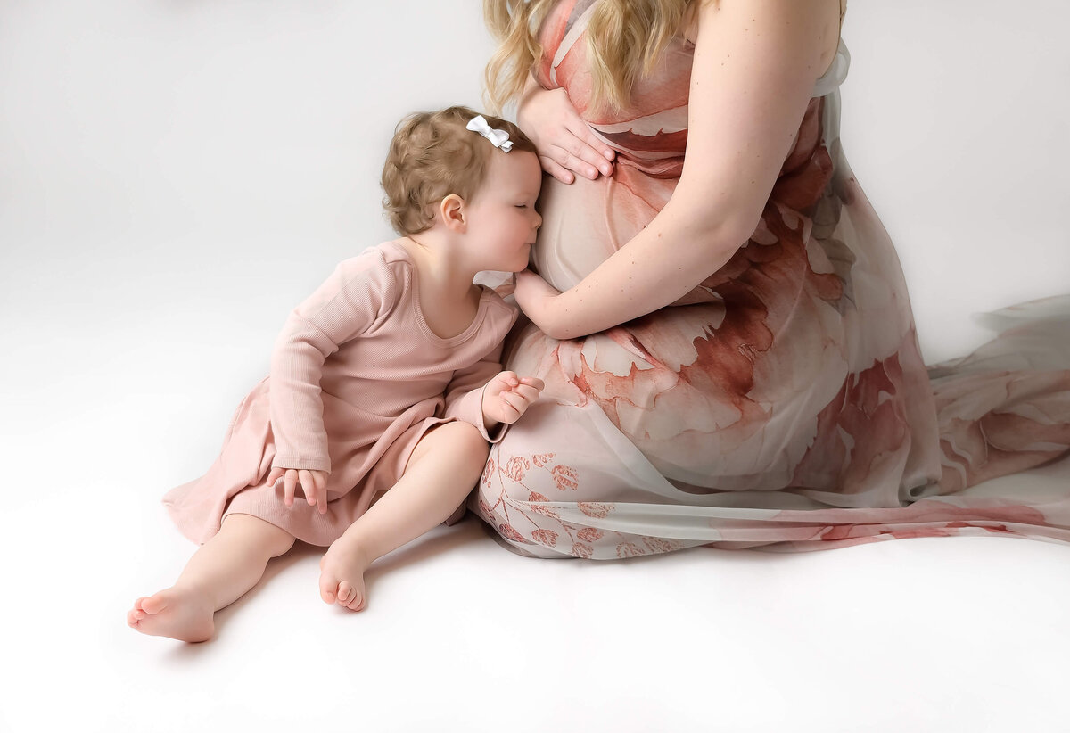 Maternity session close up of toddler kissing mom's pregnant belly as they sit on the floor together. Image by Beth Benecki.