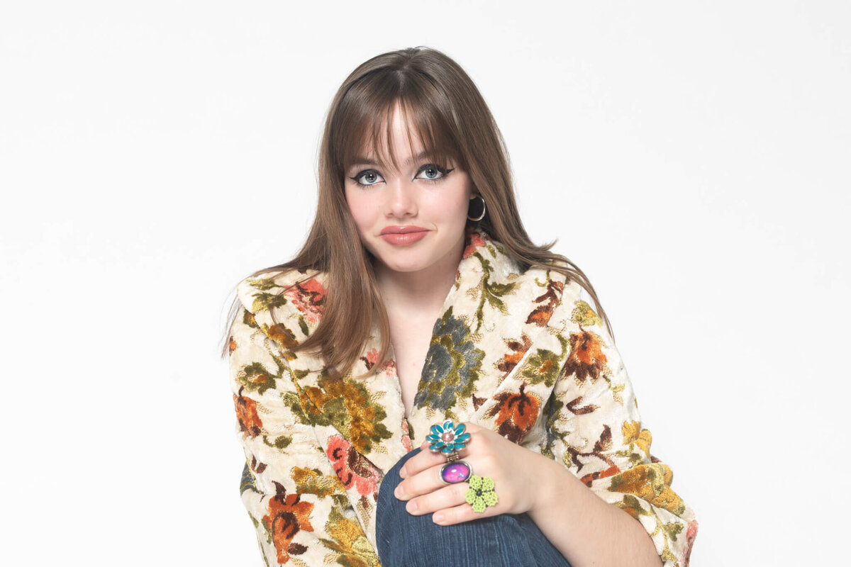 A woman with long brown hair wears a floral-patterned jacket and denim. She smiles softly, displaying large, colorful rings on her fingers. The background is plain white.