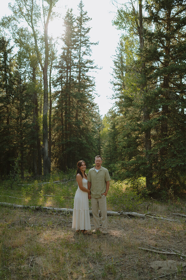 Summer forest couples session near Boise ID. A woman holds the hand of her husband as they both look at the camera and smile.