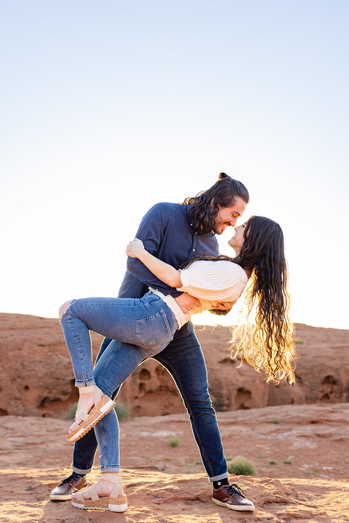 Engaged couple stares into each others eyes as he dips her back, showing off her hair in the sunshine