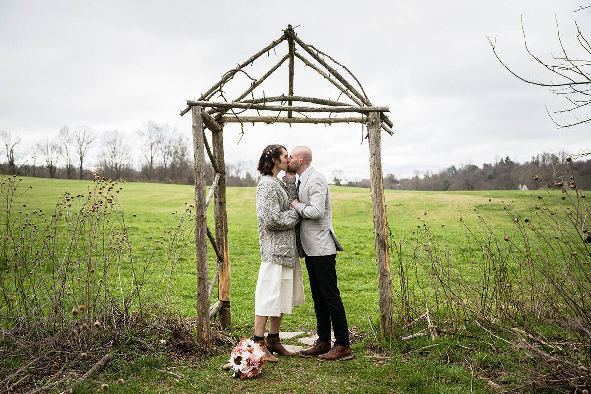 A couple stands under a structure made of logs and sticks to have their first kiss on their wedding day.