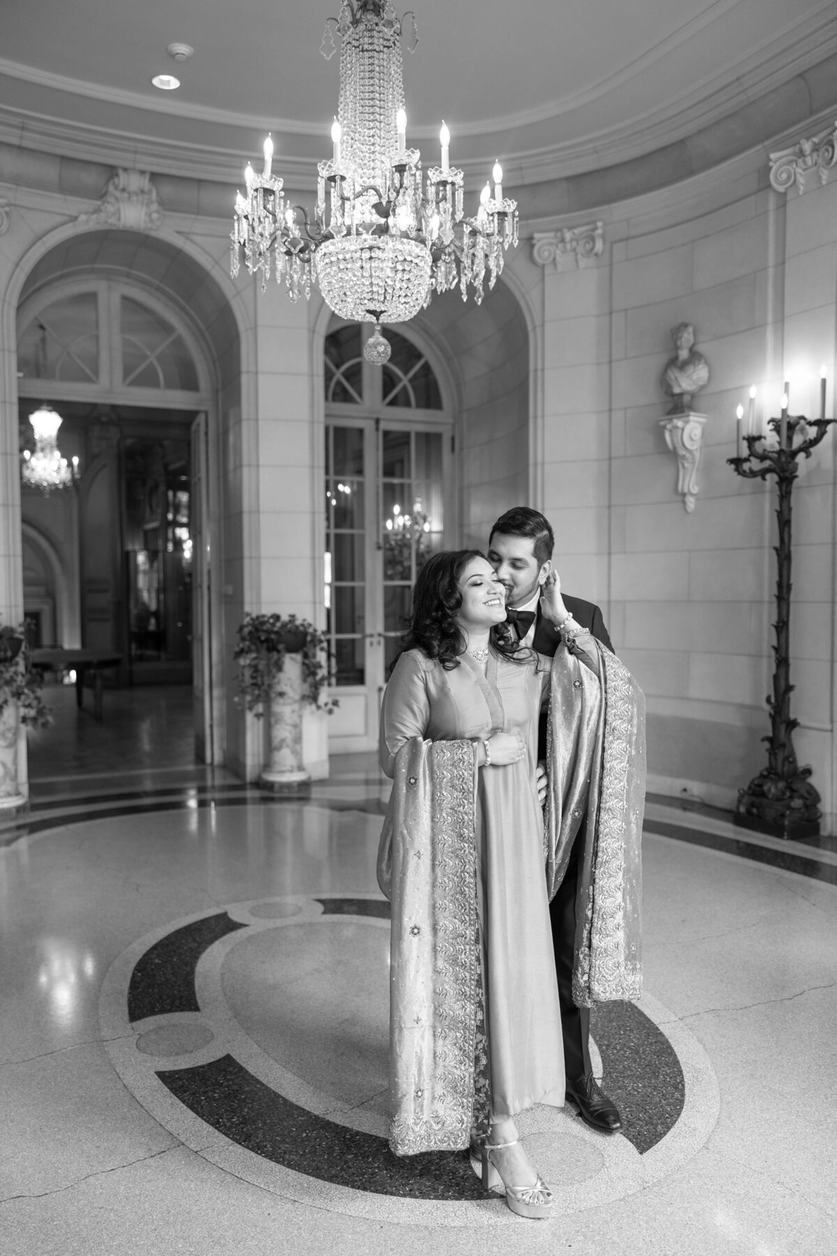 A couple stands under an ornate chandelier in an elegant room. Both are dressed in formal attire with an elaborate cloak draped over their shoulders. The background features a large doorway and classical sculptures. The image is in black and white.