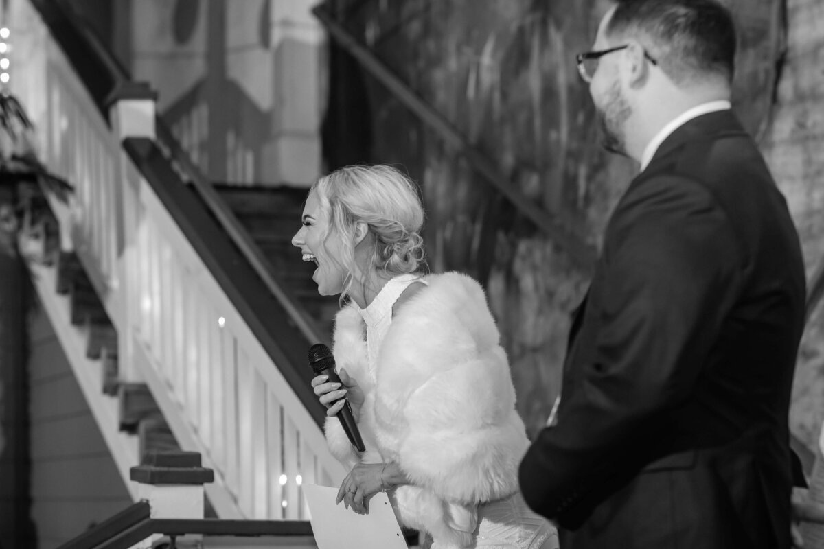A woman in a white dress and fur shawl laughs while holding a microphone and paper, standing next to a man in a suit and glasses. They are outdoors near a staircase with candles on the steps.