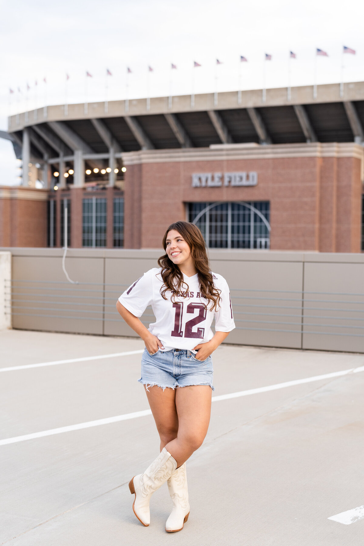 Texas A&M senior girl wearing white jersey and boots with hands in pockets and smiling away on top of a rooftop parking garage with Kyle Field in background
