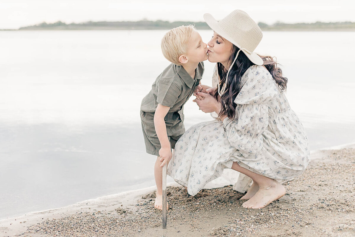 Mom kneeling on the beach in Kansas City, gently kissing her toddler son's cheek during milestone photography session.