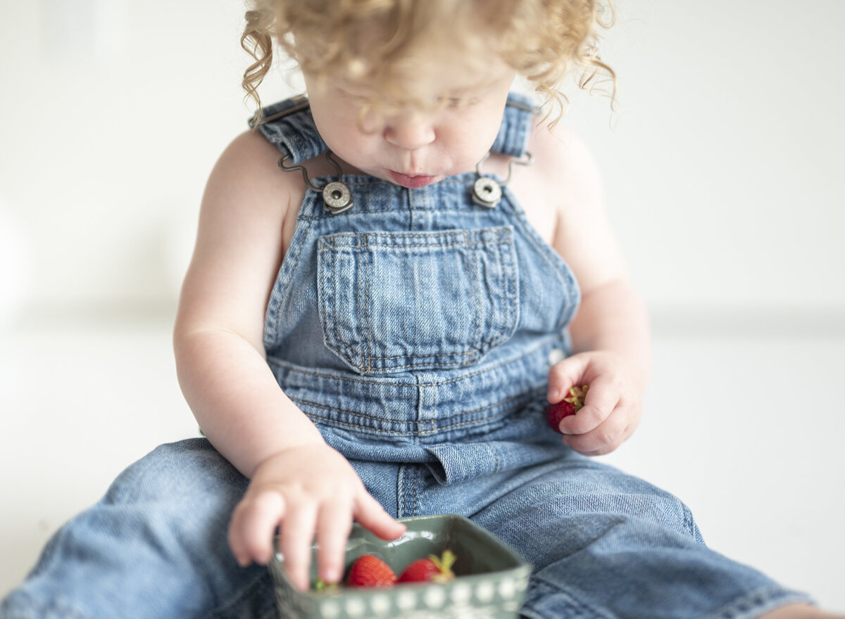 Toddler in denim overalls sitting indoors, eating strawberries.