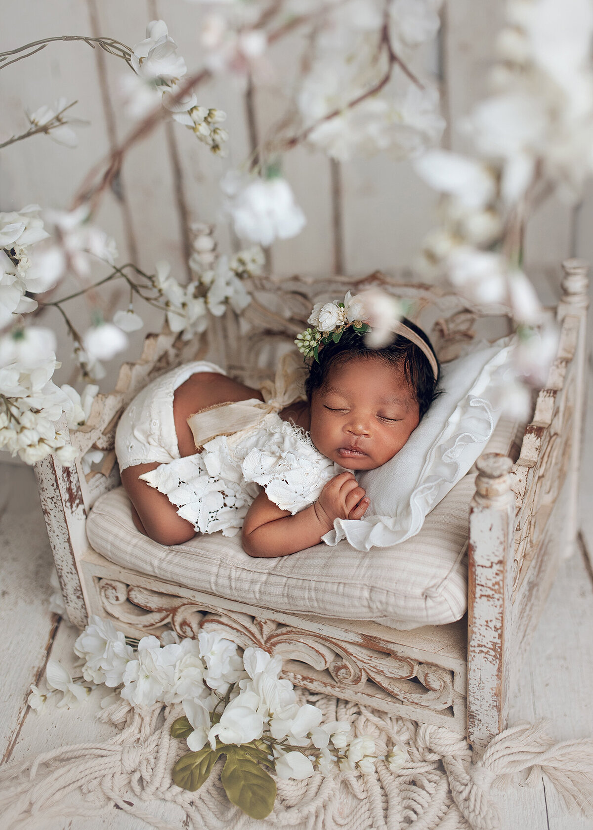 sweet newborn baby dressed in a white outfit laying on little scroll wood bed under blooming branches with white flowers