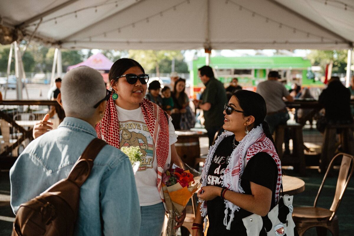 Three people with sunglasses and kufia's talking at an event.