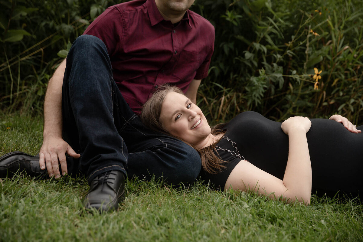 Pregnant wife in black dress laying on the grass in her husband's lap.