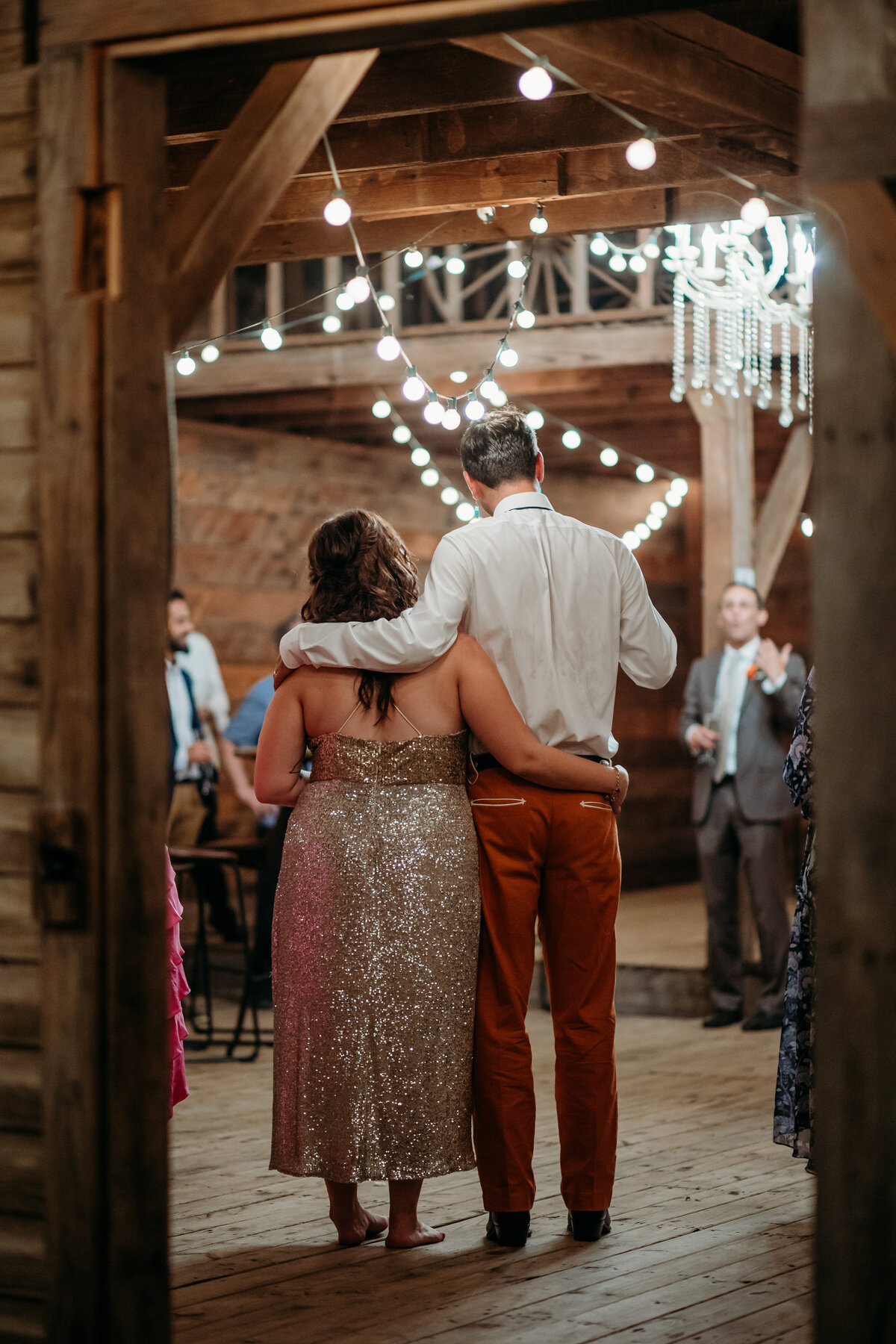 Couple walking arm in arm into a rustic barn adorned with string lights, viewed from behind