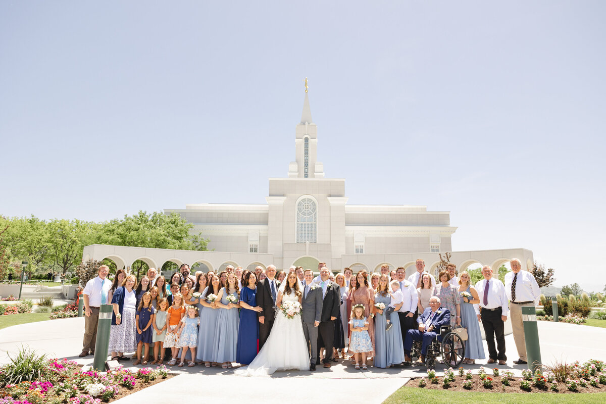 A large wedding party gathers in front of the Bountiful Utah LDS Temple, posing for a joyful group photo in the sunshine.