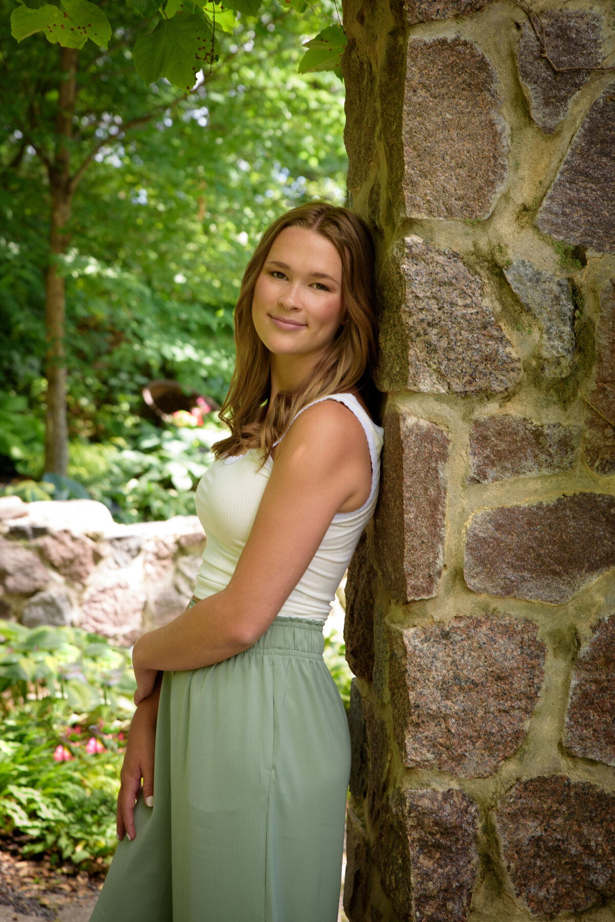 Luxemburg Casco High School senior girl wearing long sage green pants and a white top by stone building in the flower gardens at the Green Bay Botanical Gardens in Green Bay, Wisconsin