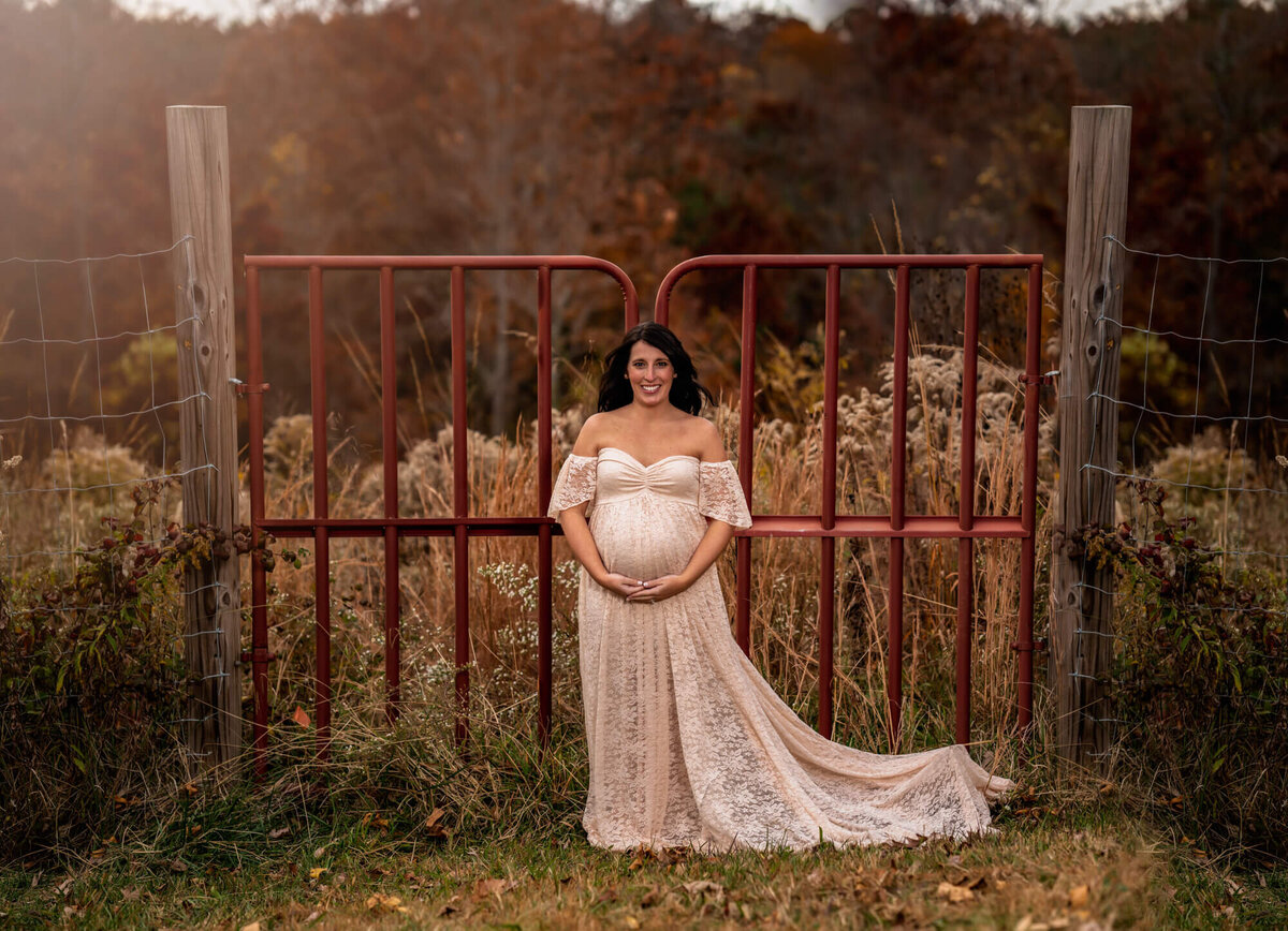 A beautiful mama to be in a white lace dress cradles her baby bump while standing in front of a red gate