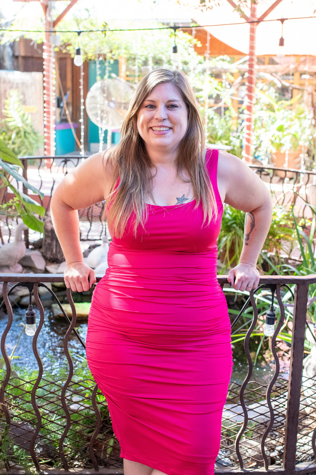 Smiling woman in a bright pink dress posing by a garden pond and railing outdoors, photographed by Vyrl Photo, showcasing Tucson brand photography.