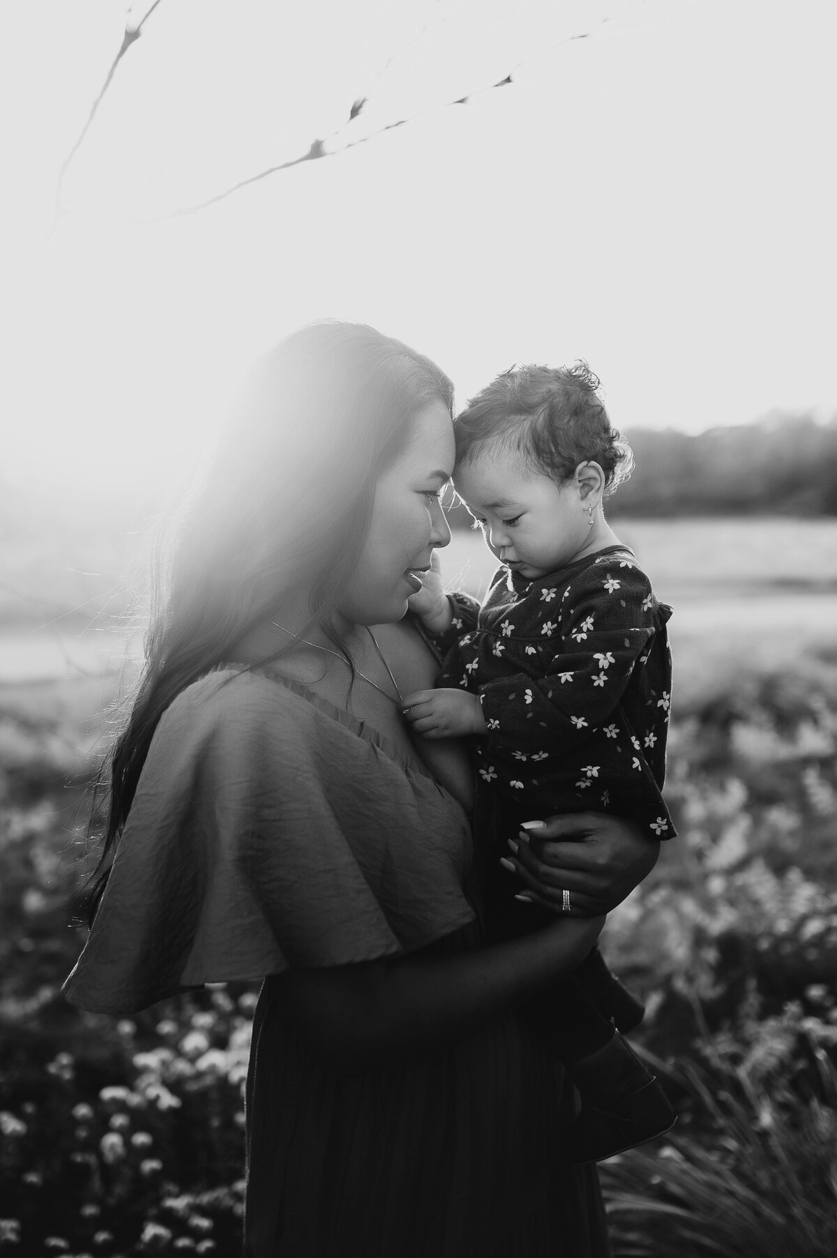 A mother holding her baby girl and pressing their foreheads together with the sun shining through. 