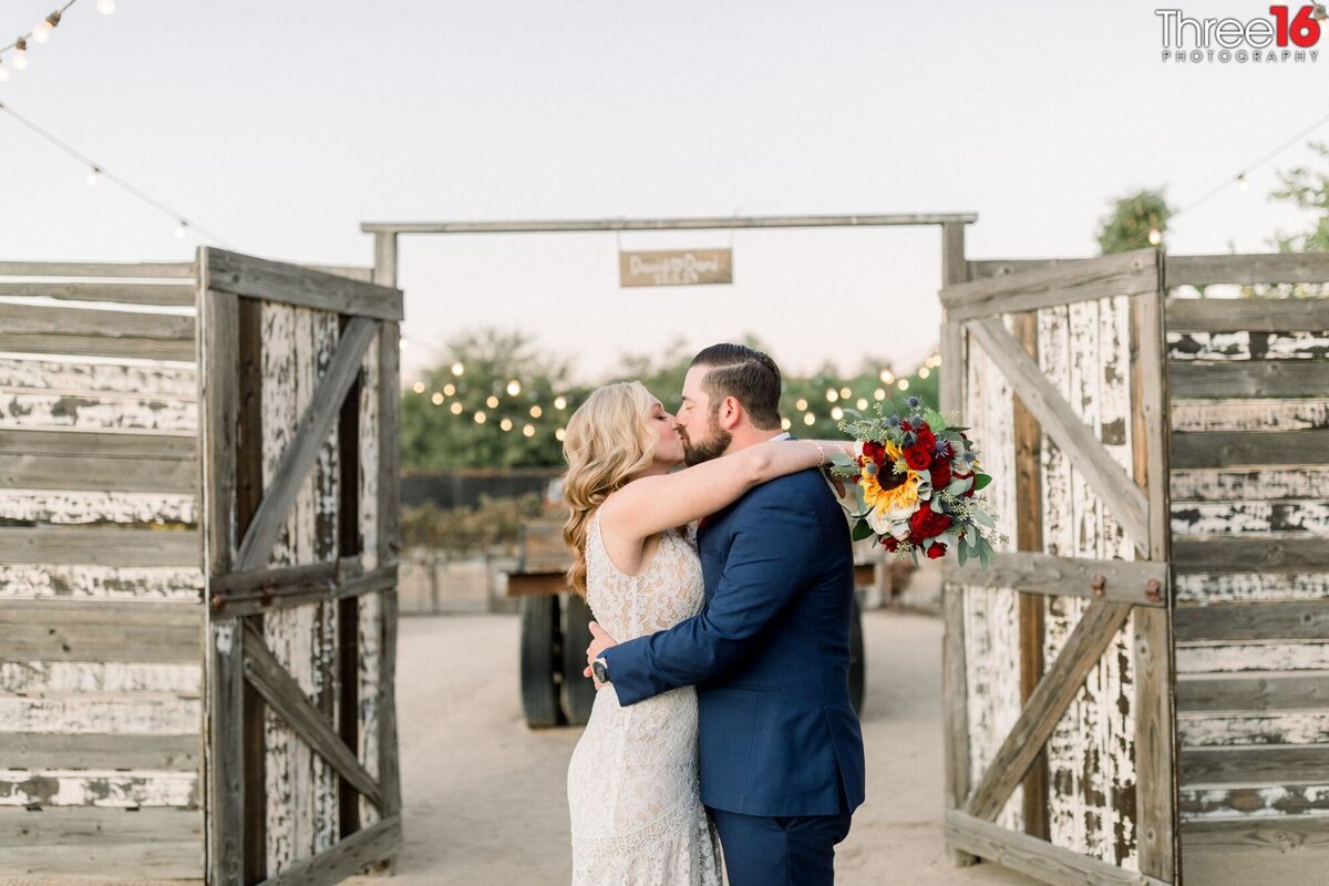 Bride and Groom fully embrace each other in a hug as they share a passionate kiss next to the rustic open gate