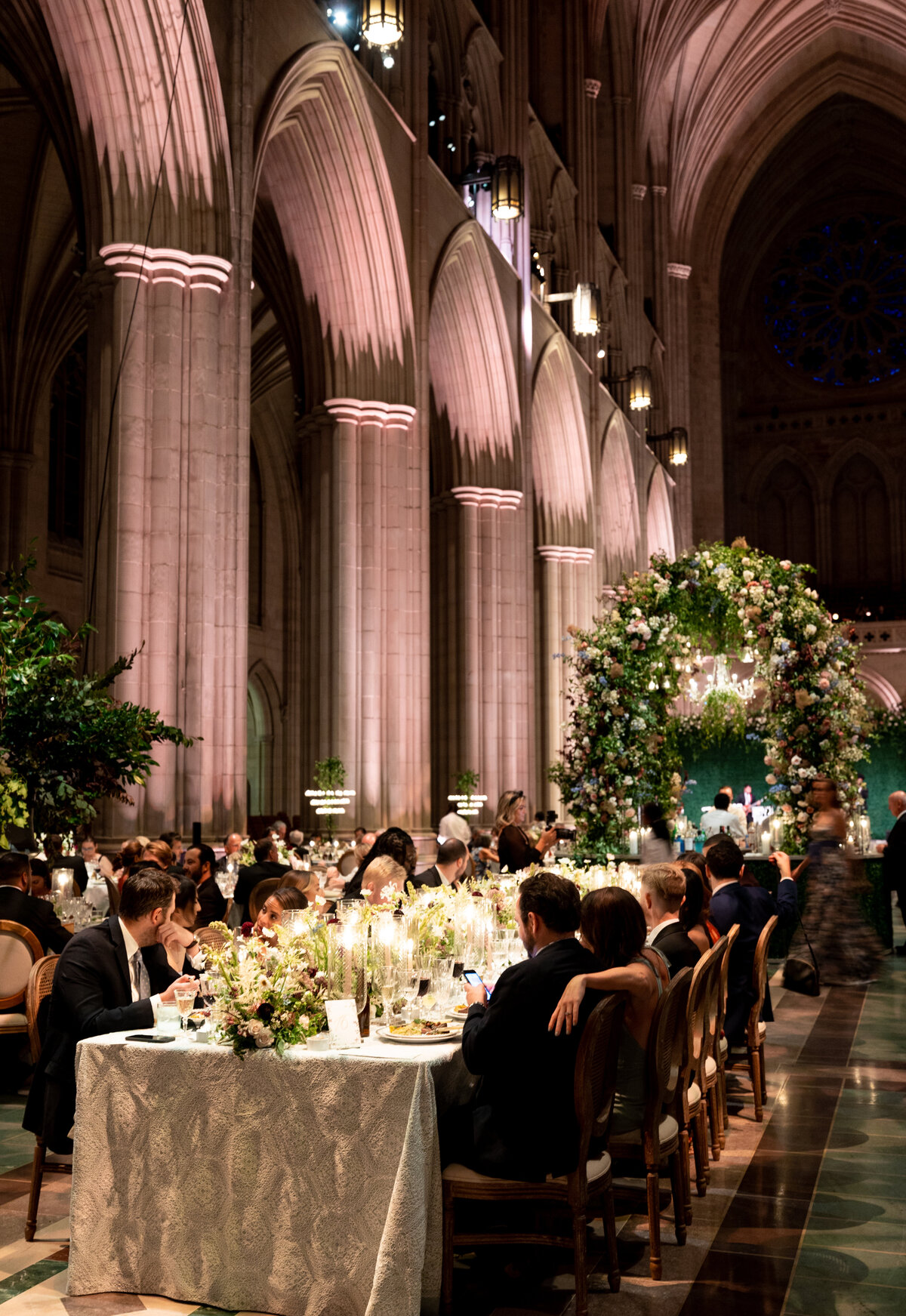 This stunning photograph was captured at Washington National Cathedral in Washington D.C. by photographer Twah Dougherty. The image showcases a grand hall with high arches adorned in greenery and flowers, creating a beautiful backdrop for a formal dinner event. Long tables are elegantly set with intricate flower arrangements and soft candlelight, as people dine and socialize in a sophisticated atmosphere. A lavishly decorated floral arch stands prominently at the far end, adding a touch of charm and elegance to the overall scene.