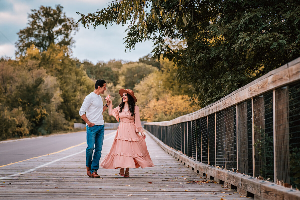 A couple walking hand in hand on a wooden bridge in a picturesque, tree-lined setting. The woman is wearing a pink dress and a hat, while the man is dressed casually in jeans and a white sweater.