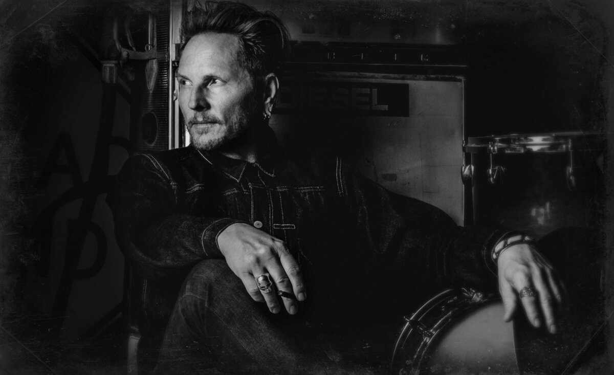 Matt Sorum portrait black and white sitting against gas pump surrounded by drums