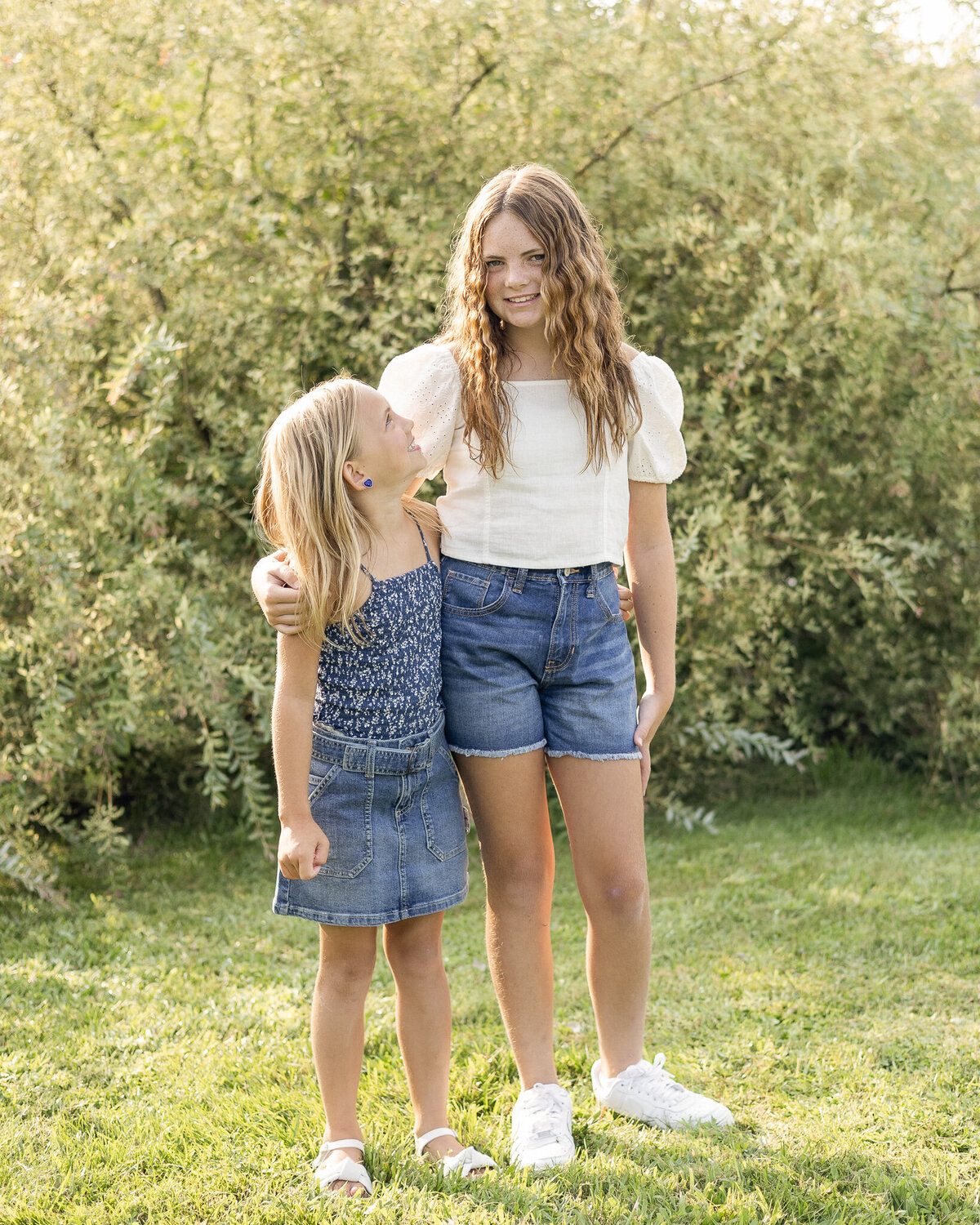 Sisters standing in the grass next to each other in front of a bush, younger sister looking at older sister smiling while older sister is looking at the camera smiling.  Older daughter is wearing blue jean shorts, a white short sleeve shirt, and has long brown hair.  Youngest daughter has a dark blue and white tank top style shirt with a blue jean skirt, and has medium length blonde hair.