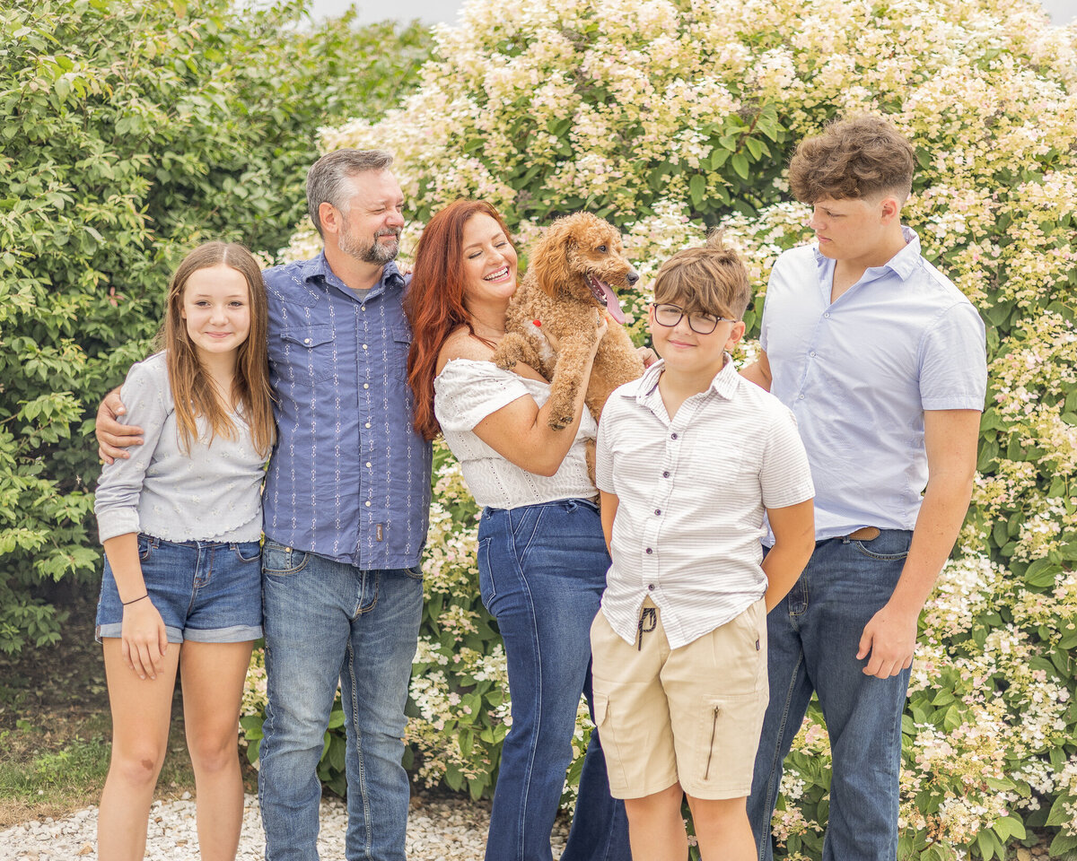 Family of five plus their light brown dog are standing in front of a bush. Some are looking at the dog smiling and others at the camera smiling.  Daughter is wearing blue jean shorts, a long sleeve light blue shirt, white shoes, and has long light brown hair.  Dad is wearing blue jeans, blue and white stripped button up shirt, and has short pepper hair and beard.  Mom is wearing blue jeans, short sleeve white shirt, and has long red hair.  Younger son is wearing a light tan button up and khaki shorts, white shoes, short light brown hair.  Oldest son is wearing a light blue short sleeve button up, blue jeans, and has short light brown hair