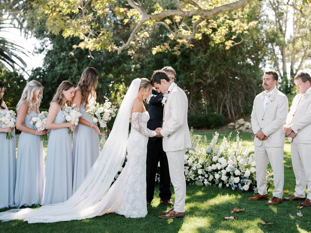 The wedding party surround Lily & Mitchel as they say I do. Primrose & Petal's bouquets line the ground at the alter with white flowers and baby blue accents.