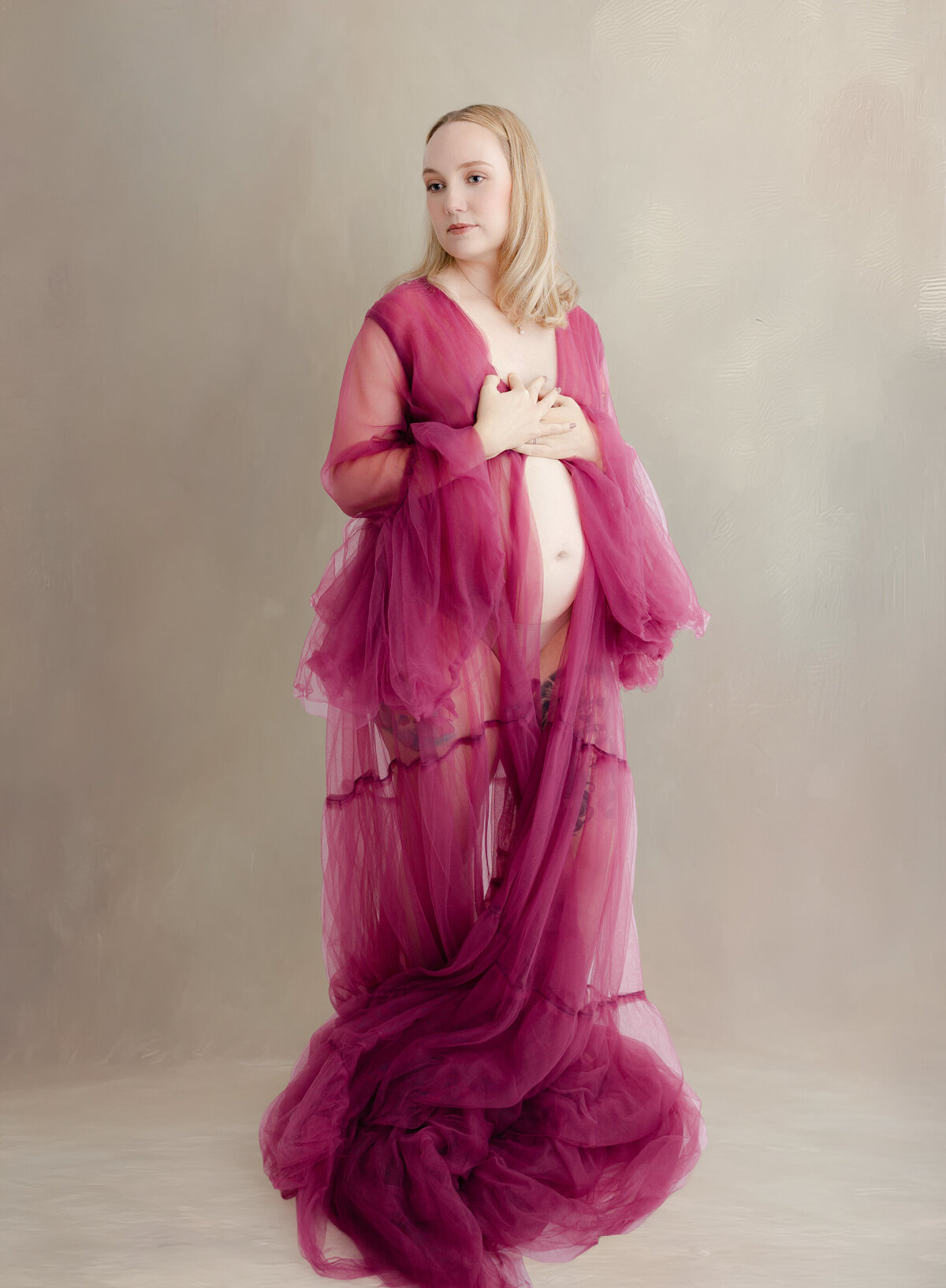 Pregnant mom wearing a deep pink robe and posed with belly showing against a light brown backdrop.