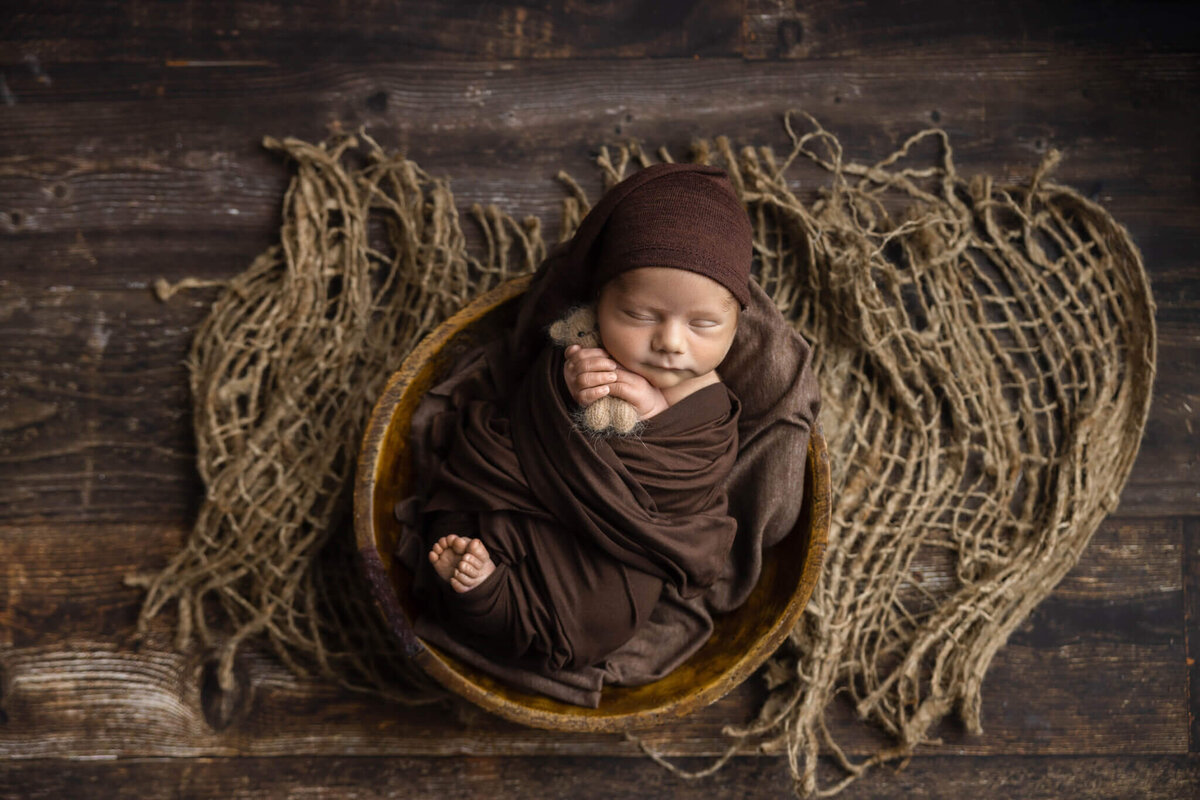 newborn baby wrapped in a brown fabric holding a tiny teddy bear asleep in a prop bowl