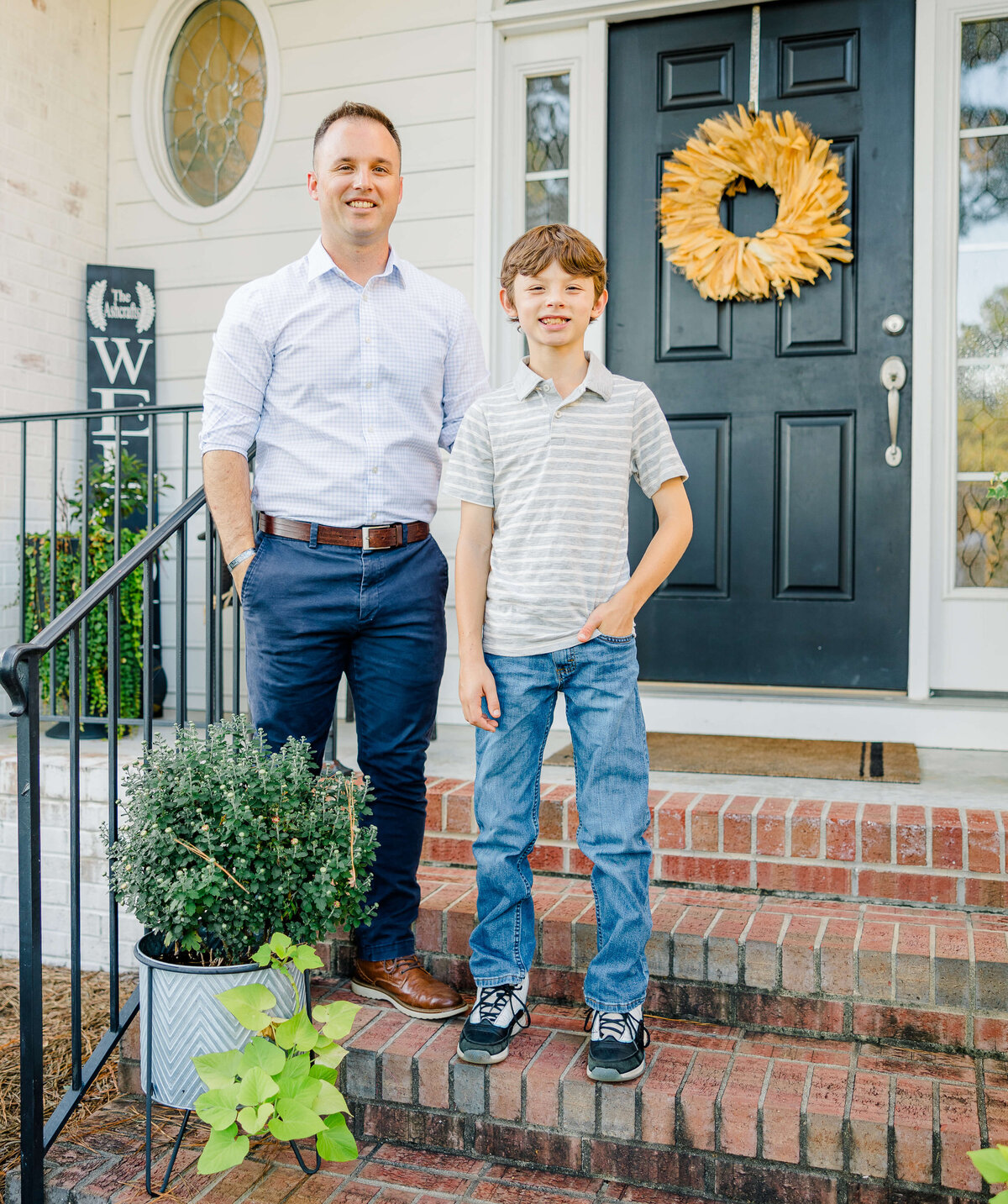 A man and a boy stand on the steps of a house with a black door, captured perfectly by a Fayetteville NC family photographer. The man wears a striped shirt and blue pants, while the boy matches in jeans. A plant in a pot and a wreath on the door add charm to this heartfelt scene.