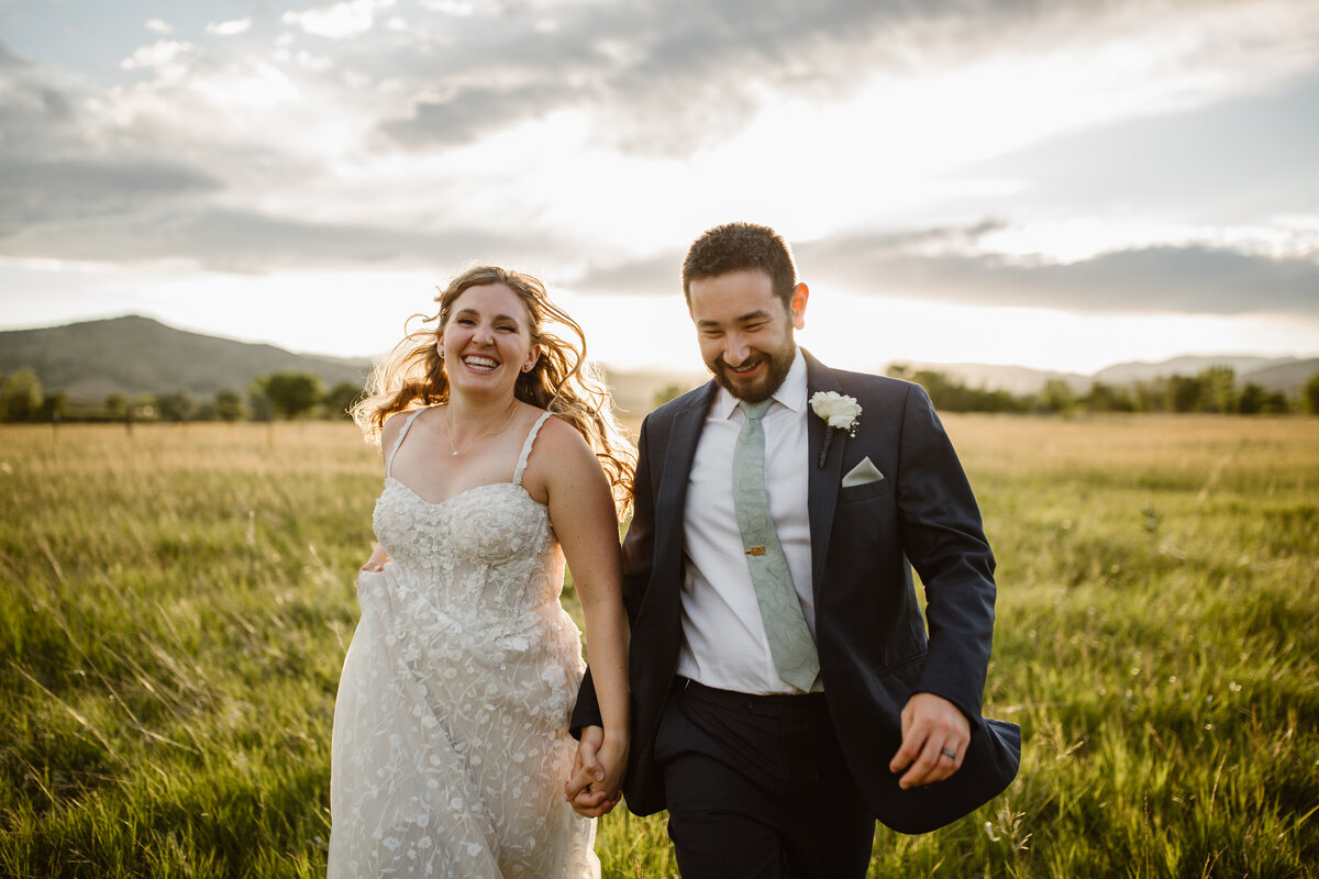 Bride and groom joyfully run hand-in-hand through a sunlit field during golden hour, laughing together on their wedding day.