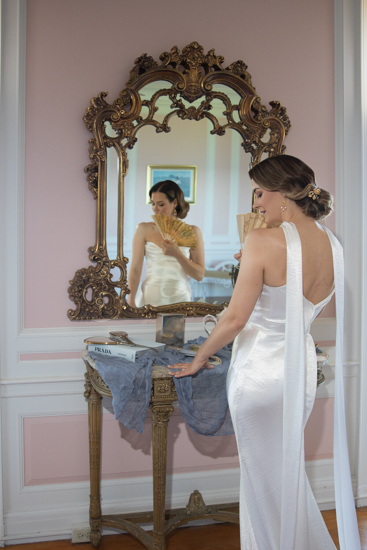 Aldrich Mansion Wedding Photos | Kelsey Sheehan Photography Timeless Rhode Island Weddings | A woman in an elegant white gown admires herself in an ornate mirror, holding a fan. The setting exudes a vintage, sophisticated atmosphere.