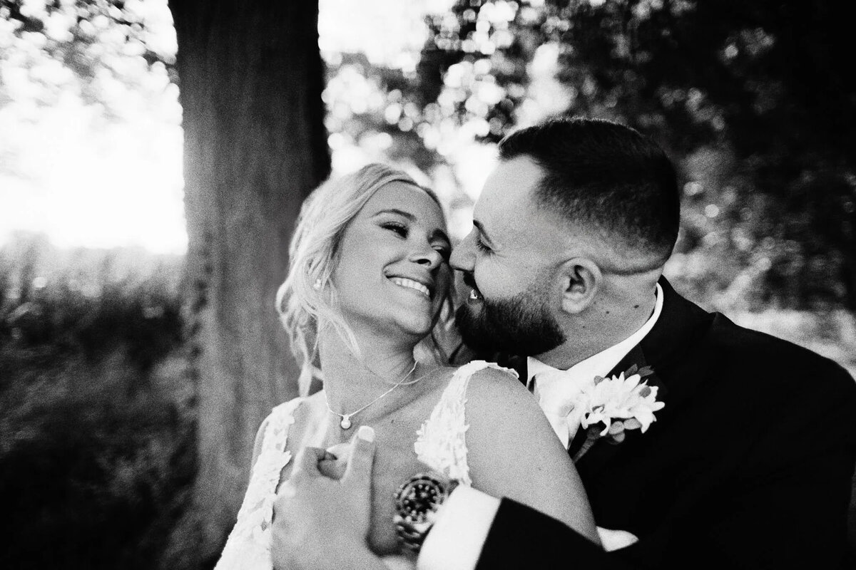 A bride and groom sit close together outdoors, smiling and gazing into each other's eyes, sharing a joyful, intimate moment on their wedding day—captured beautifully by an NJ wedding photographer. The background is softly blurred with trees.