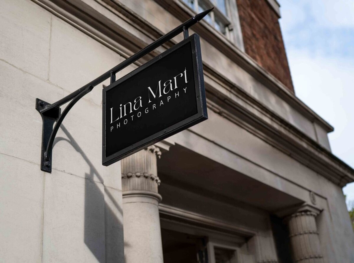 Black hanging shop sign for “Lina Mart Photography” mounted on the exterior of a classic stone building with columns.