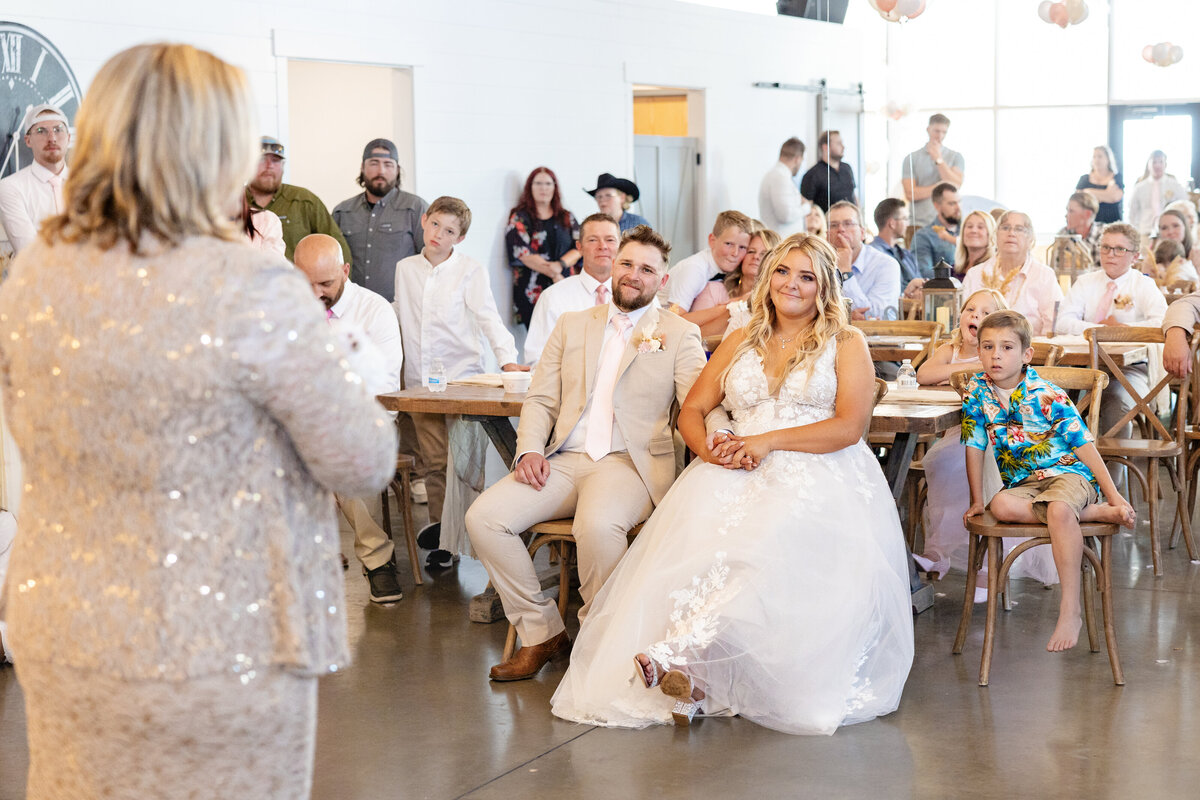 The bride and groom sit together, smiling and listening to a heartfelt speech at their wedding reception.