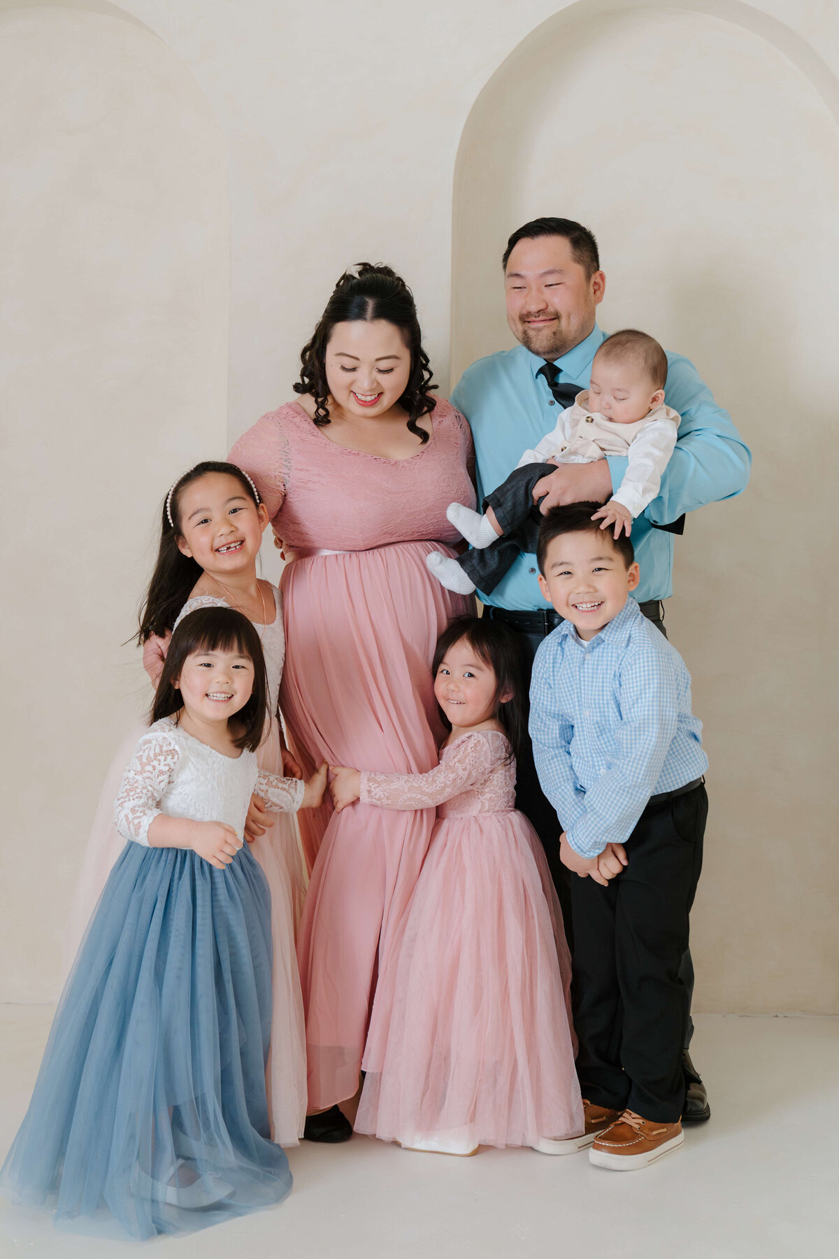 A large family huddles close, smiling and cuddling warmly in a cream-toned studio setting.