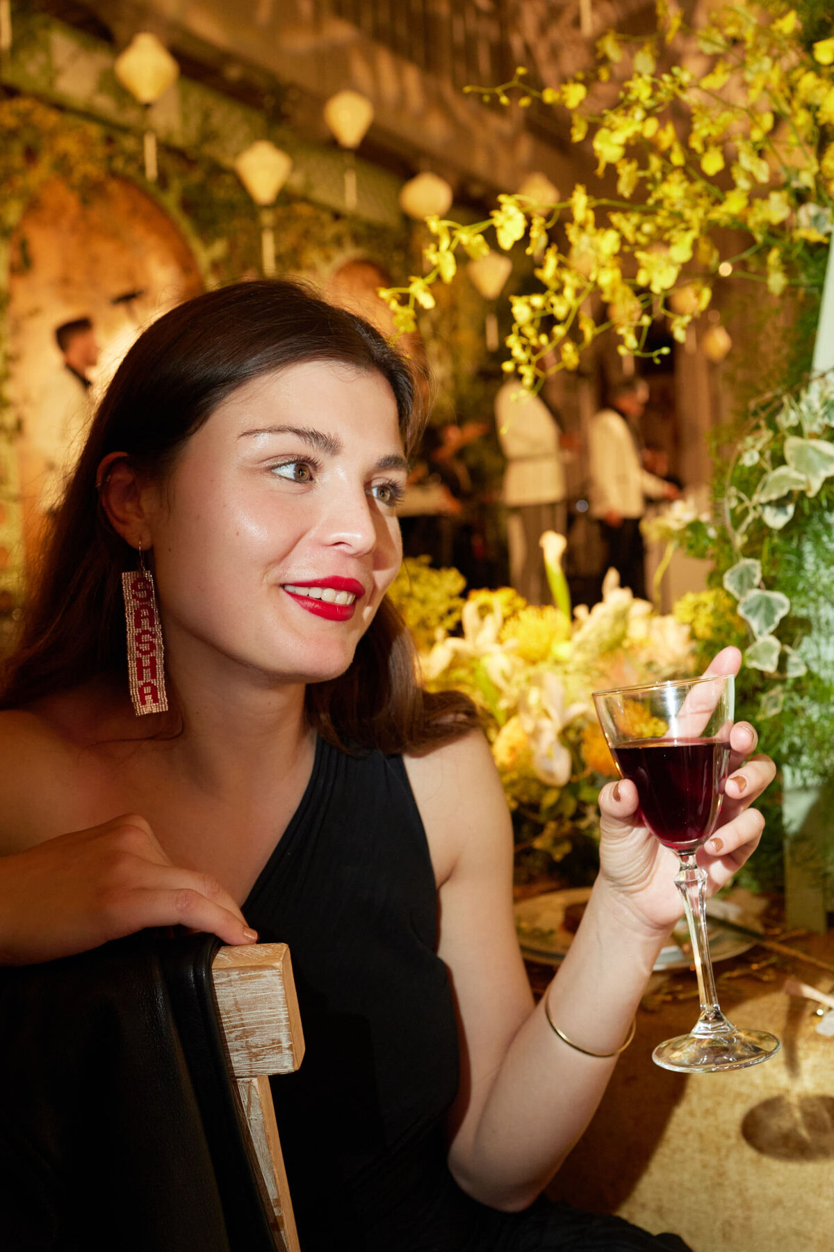 The photo was taken at The New York Public Library by photographer Shawn Connell. In the foreground, a woman with long dark hair and red lipstick, wearing a black dress and large decorative earrings, holds a glass of red wine. She is seated in a warmly lit, plant-filled indoor setting with yellow flowers at The New York Public Library. In the background, people can be seen engaged in conversation, adding to the lively atmosphere of the scene. Aaron Navak Films also provided videography services for this occasion.