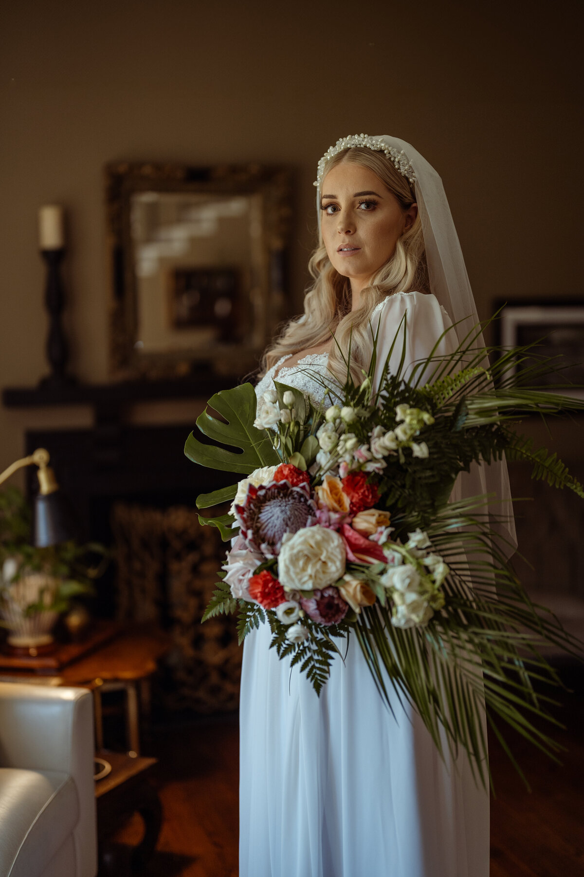 Elegant portrait of a bride moments before her ceremony, dressed in a vintage-style gown and holding a vibrant tropical bouquet. Captured in a warmly lit room, this DFW-based wedding portrait showcases the bride's classic beauty and unique floral arrangement, highlighting the intimate and sophisticated atmosphere.