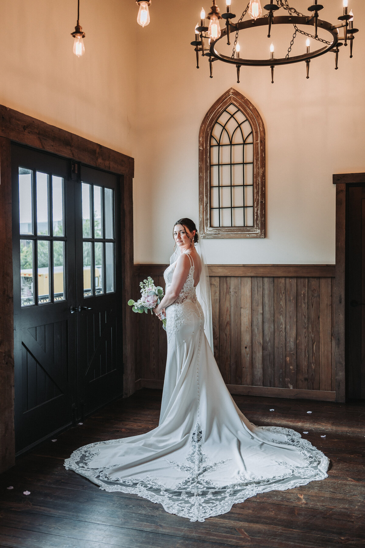 A beautiful bride in a long, flowing lace wedding gown stands elegantly in the rustic interior of Sweeney Barn in Manassas, Virginia, holding her bouquet and looking over her shoulder, captured by Quinn Photography LLC.
