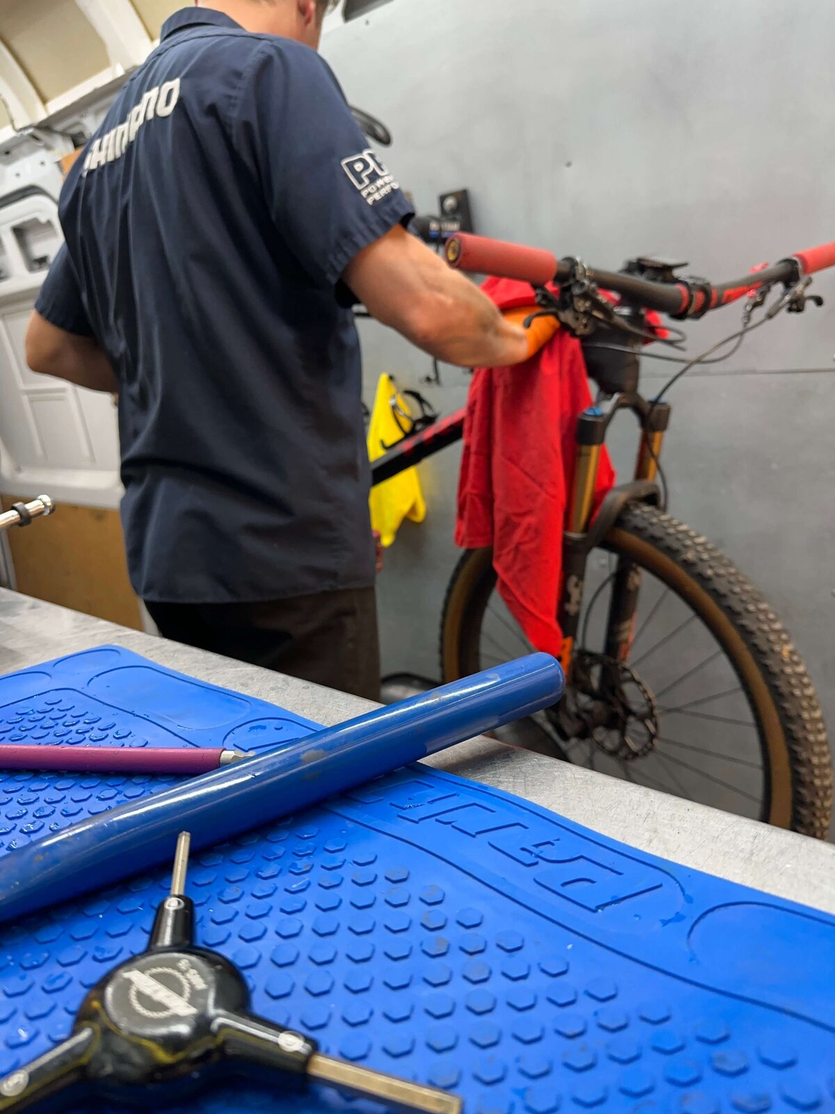 A person in a blue shirt is cleaning a mountain bike with a red cloth. The bike is supported on a stand in a workshop. Various tools are visible on the table in the foreground