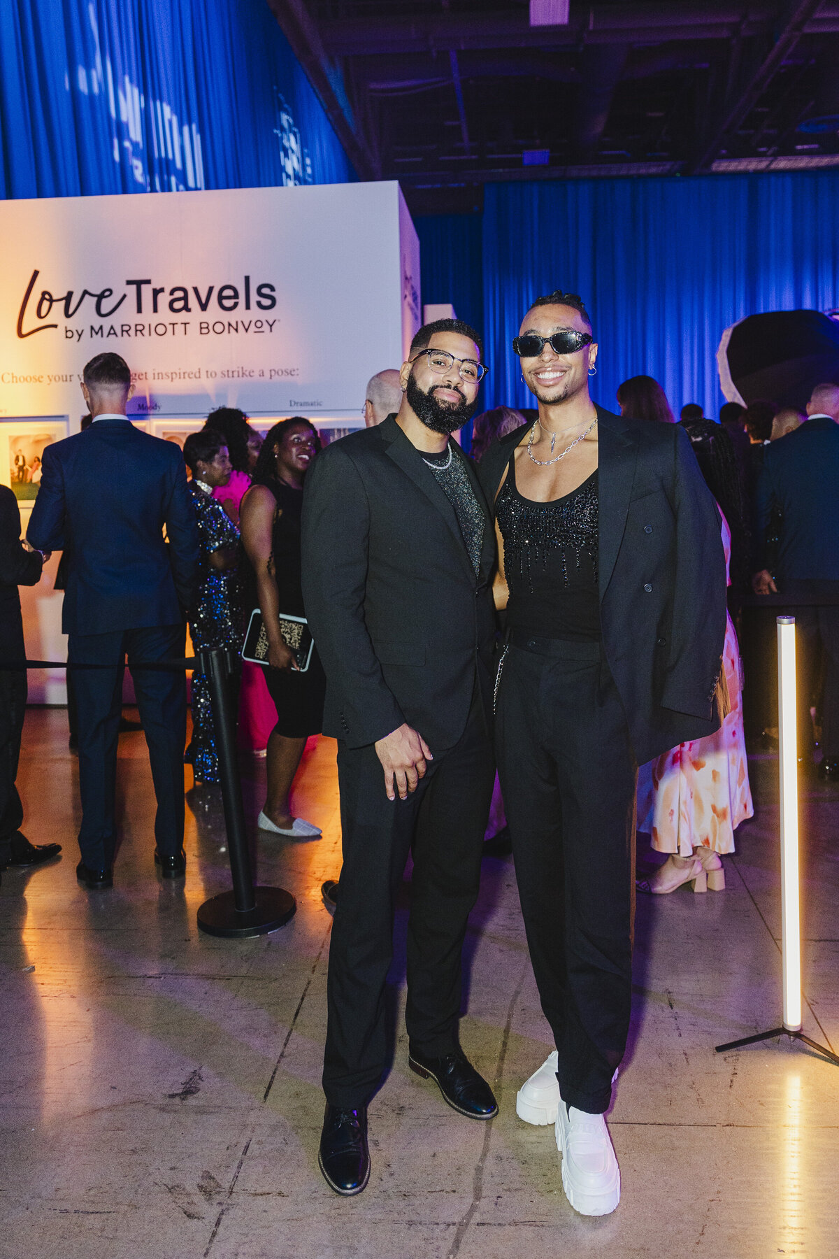 Two people pose in all black outfits with a sign behind them that says Love Travels.