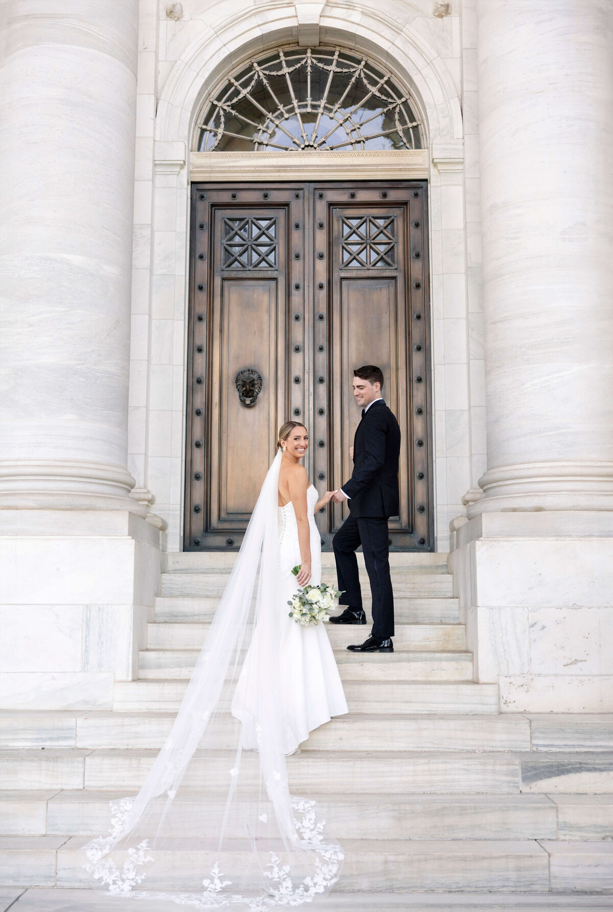 A bride and groom stand on elegant stone steps in front of a large, ornate wooden door. The bride, in a long white gown with a flowing veil, holds a bouquet. The groom, in a black suit, looks back at her. They appear happy and poised.