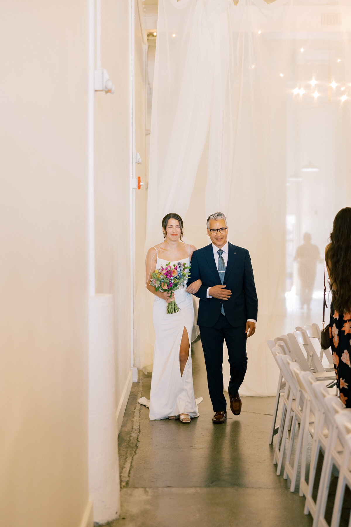 Sara in lace gown holding bouquet by Crimson & Clover walking down aisle at Fetewell wedding ceremony.