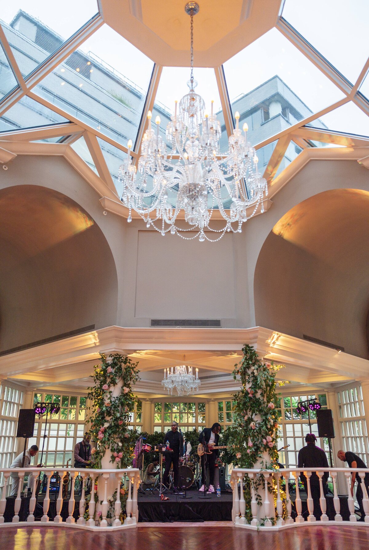 A spacious, elegant ballroom with a high glass ceiling features a large chandelier. A band performs on a stage surrounded by floral arrangements and soft lighting, creating a festive atmosphere.