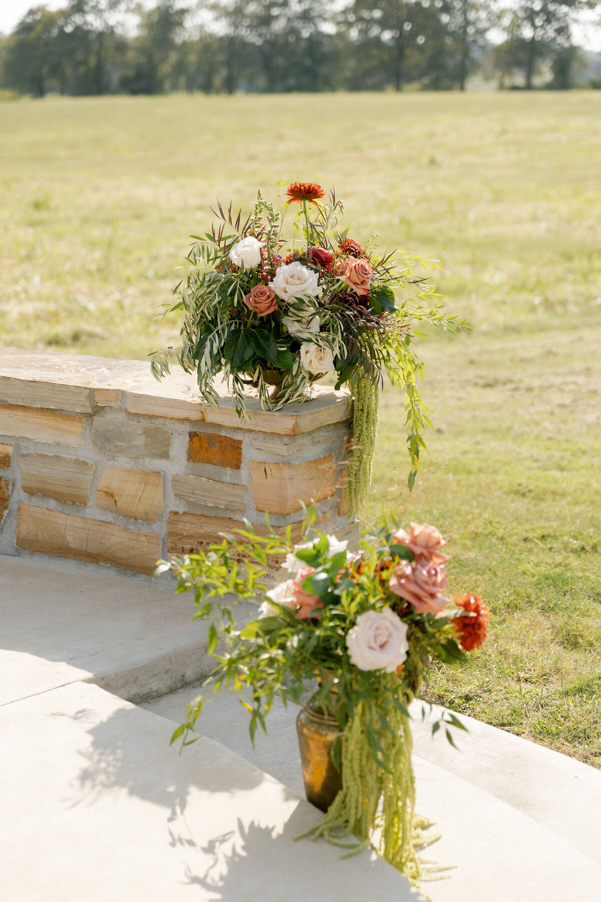 Two garden-style wedding floral arrangements rest on and beside a stone wall, showcasing a palette of cream, blush, peach, rust, burgundy, and vibrant greenery. Trailing amaranthus and mixed foliage add movement and texture. The ceremony backdrop features open grassy fields and distant trees under soft sunlight at a rustic-modern outdoor venue.