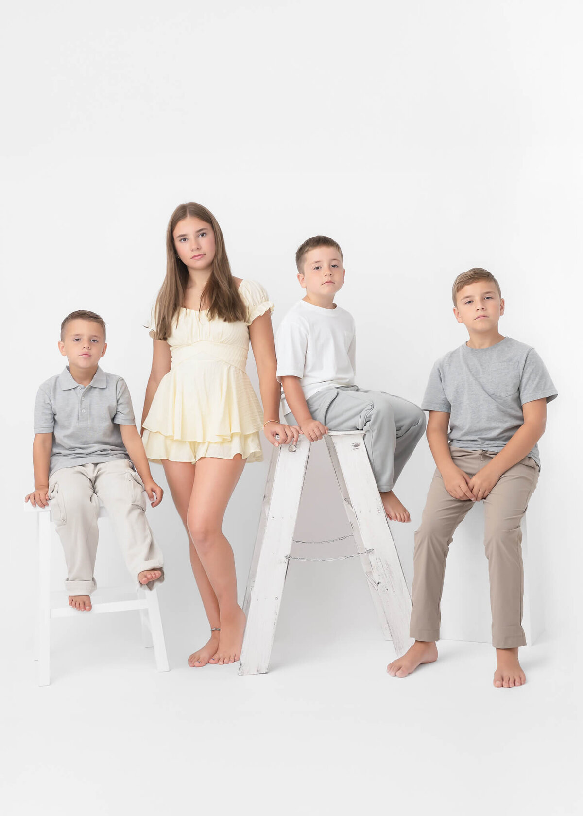 4 siblings, 3 younger boys one teen girl posed in serious face portrait in white studio wearing neutrals, boys sitting, one on ladder girl standing in yellow dress