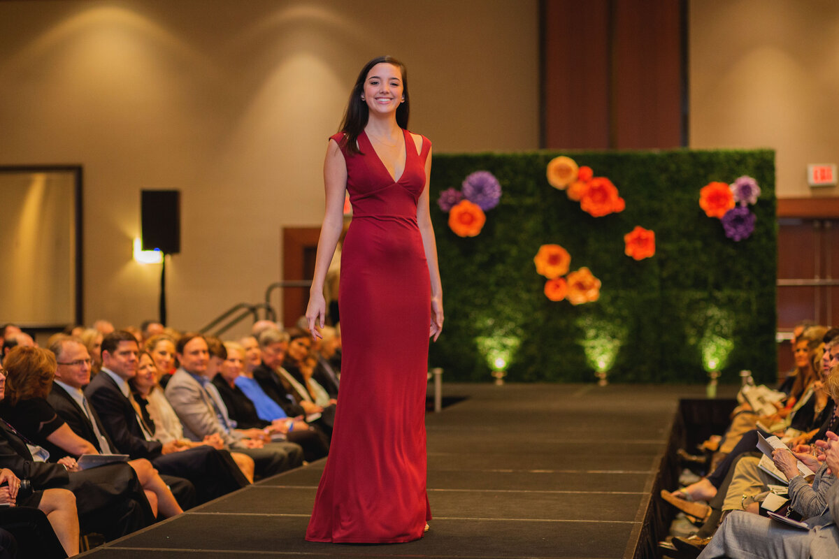 Model in a red dress walking on a fashion show runway with a paper flower backdrop display