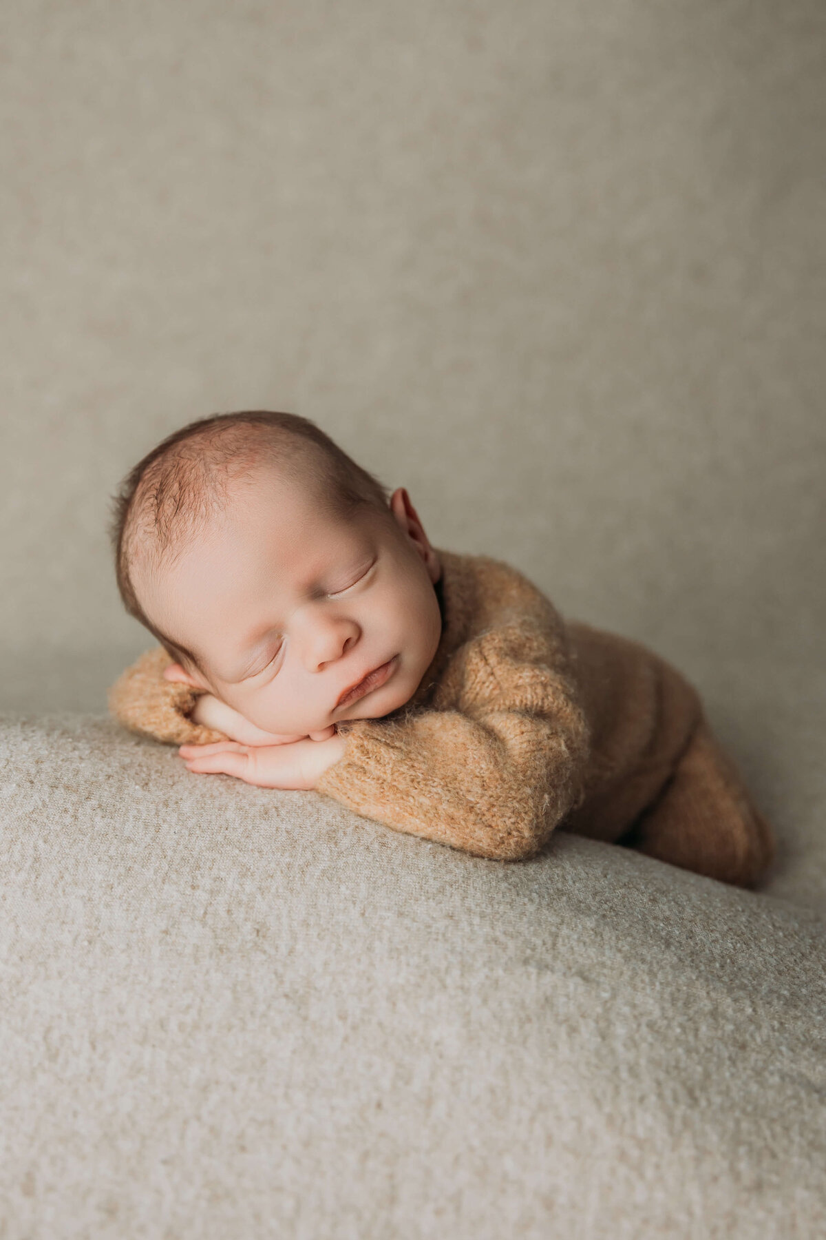A serene newborn baby rests on a soft, textured surface, with their head resting on folded arms, eyes closed, and a slight smile on their face. The baby wears a cozy, neutral-colored outfit, and the background is softly lit with a neutral tone.